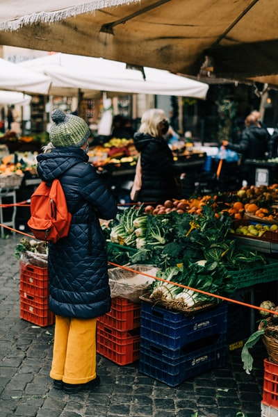 一位戴着黑色针织帽、身穿橙色外套的妇女站在绿色蔬菜展台前